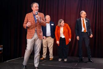 UT Austin President Jay Hartzell on stage holding mic with three people standing behind