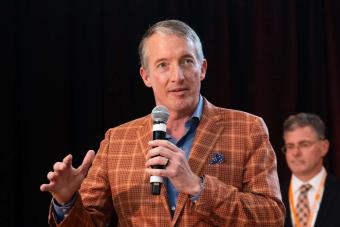 UT Austin President Jay Hartzell on stage holding mic with a person in the background