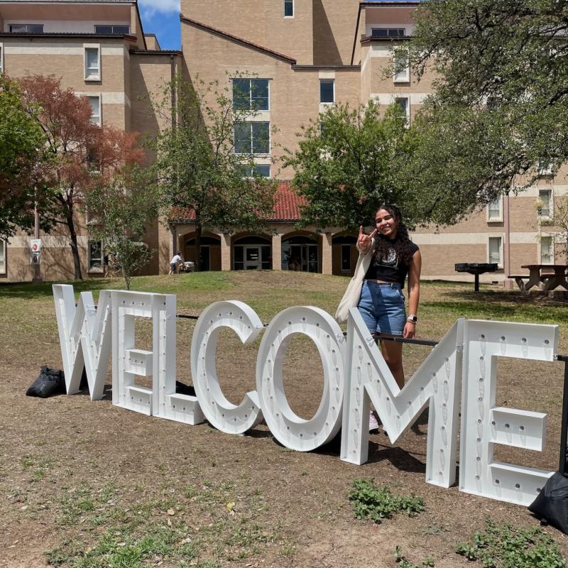 Student standing behind welcome sign flashing hook-em hand sign