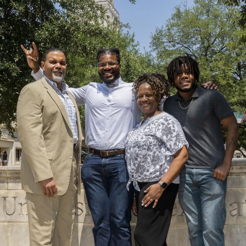 Four family members standing together and smiling with UT tower behind trees in background