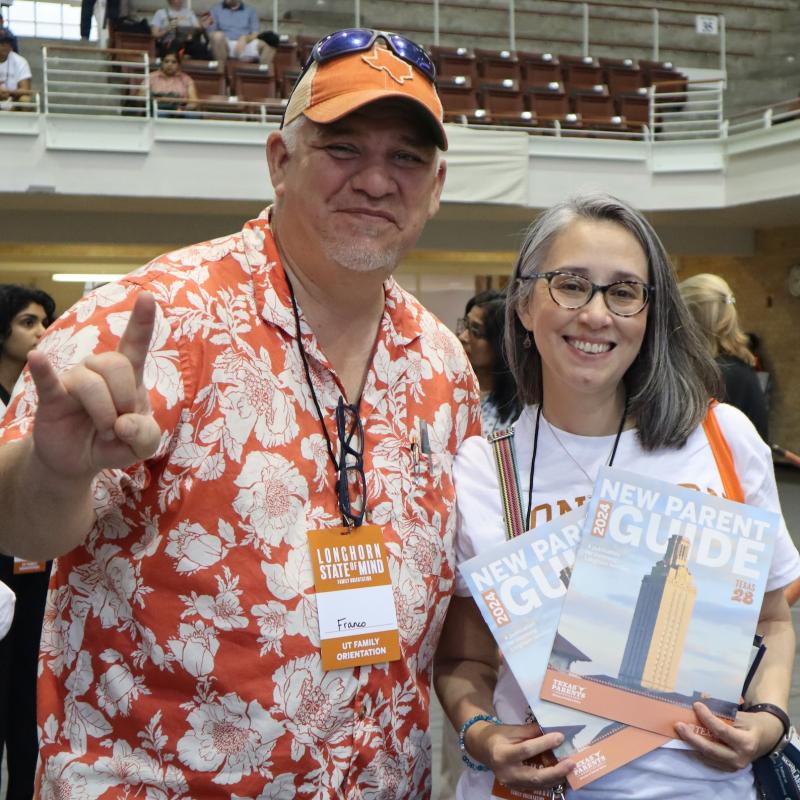 Couple holding UT Austin’s New Parent Guide and flashing Hook ‘em hand sign